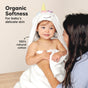 A smiling toddler wrapped in a white unicorn hooded towel sits on a wooden surface, facing a woman. Text reads, Organic Softness for babys delicate skin and 100% natural cotton.