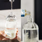 A hand fills a Grove Collaborative Tub & Tile Cleaner Starter Set glass spray bottle under a faucet while tub and tile cleaner is poured into another clear bottle nearby.