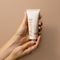 A pair of hands gently cradles a tube of PAUME Probiotic Hand Balm against a beige backdrop. The tube, with natural probiotics and essential oils, features light hues with slightly darker text, enhancing the serene aesthetic.