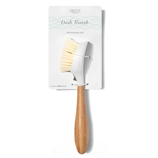 A dish brush with a wooden handle and white bristles is attached to a cardboard backing labeled “Grove Dish Brush with Replaceable Head.”.