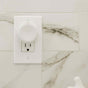 A white electrical outlet with a round white safety cover plugged into the top socket, mounted on a wall with white and gray marbled tiles.