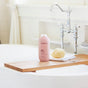 A bottle of method Body Wash with a cruelty-free formula rests on a wooden bath tray beside a loofah and folded towel in a clean, white bathroom featuring a classic silver faucet above the freestanding tub.
