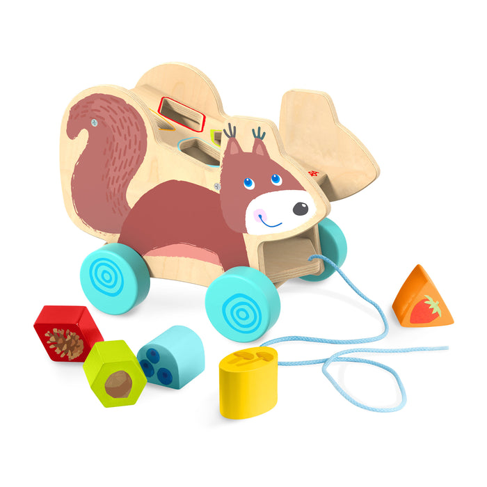 Wooden pull-along toy shaped like a squirrel with blue wheels, featuring shape-sorting slots on its back. Colorful geometric blocks are scattered nearby, including red, yellow, blue, and orange pieces.