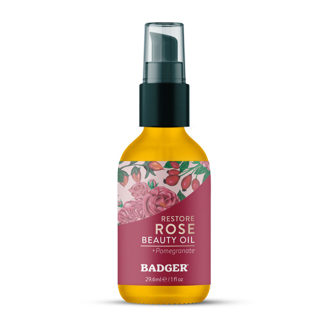 A 29.6 ml (1 fl oz) bottle of Badger Balm Rose Beauty Oil - Restores & Smooths, featuring a floral and pomegranate label design with pink accents and a black pump cap. Infused with organic rose oil to enhance skin hydration.