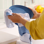 In a kitchen, a person uses a Grove Co. Organic Cotton Striped Kitchen Towel to dry a blue plate. The countertop features essentials like a sink and blurred fruit bowl with lemons in the background.