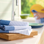 A stack of Grove Co. Organic Cotton Striped Kitchen Towels in blue and white rests on a wooden cutting board. In the background, someone uses another quick-drying towel to dry a yellow plate by the window, revealing a lush green view outside.