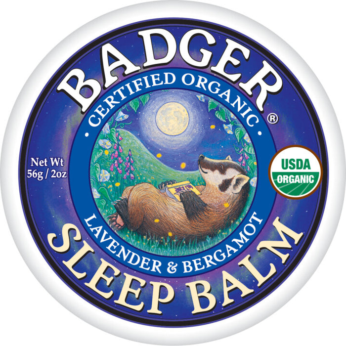 A round tin label for Badger Sleep Balm shows a badger lying on its back under a full moon, surrounded by flowers and hills. The text reads “Badger Sleep Balm,” “Certified Organic,” and “Lavender & Bergamot.”.