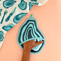 A hand slips into The Neighborgoods Oyster Pot Holder, featuring a blue and black agate-inspired design. Next to it lies a patterned cotton towel with utensil and stone motifs—an eye-catching kitchen essential.