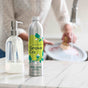 A bottle of Grove Co. dish soap stands on a kitchen counter next to a soap dispenser. In the background, a person washes dishes with a brush in a sink filled with soapy water.