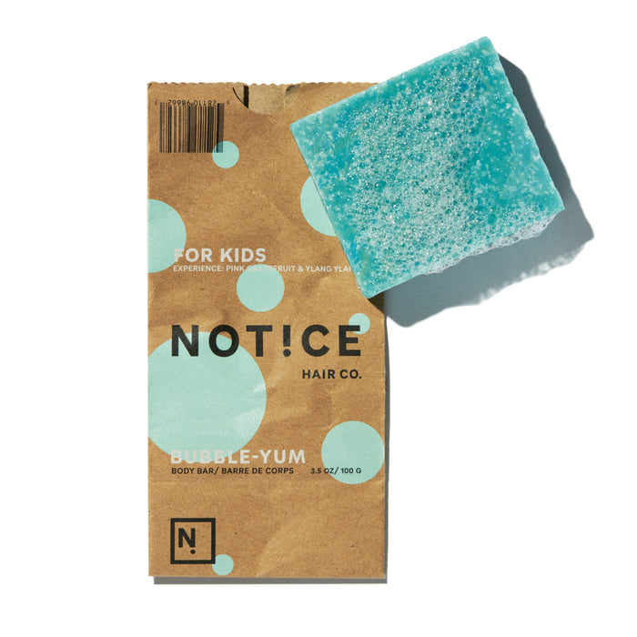 A rectangular blue bar of soap sits on a brown paper wrapper labeled Notice Hair Co. Bubble-Yum Body Bar for Kids, decorated with light blue bubbles.