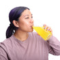 A woman with purple hair in a ponytail, wearing a mauve top, is holding and drinking from a large clear glass filled with yellow liquid against a white background.