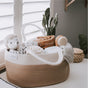 A woven basket filled with baby items, including a plush toy, rolled blankets, a baby brush, and a swaddle, sits on a white surface near a window with shutters. A potted cactus and a lidded wicker basket are nearby.