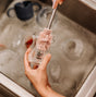 A person carefully cleans a glass baby bottle with the pink Double-Ended Silicone Cleaning & Bottle Brush from Haakaa over a sink full of soapy water and dishes. The bottle features a logo with a small heart and spiral design, highlighting the importance of hygiene in baby gear cleaning.