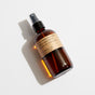 A brown glass spray bottle with a beige label reading Sandalwood Rose Room & Linen Spray lying on a white surface, casting a shadow.