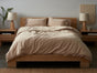 A neatly made bed with breathable beige bedding and two pillows—one featuring the Coyuchi Organic Crinkled Percale™ Sham—flanked by matching wooden nightstands with lamps and minimal decor, set against a neutral wall.