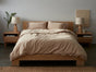 A neatly made bed features Coyuchi’s Organic Crinkled Percale™ Sheet Set in beige, crafted from GOTS certified organic cotton. The bed is centered between two wooden nightstands, each topped with a lamp; a small plant and decor sit on the left while a mug rests on the right. The room showcases breathable bedding and exudes a cozy, minimalist style.