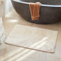 A Shag Organic Bath Rug by Coyuchi in beige rests on the bathroom floor before a large gray bathtub, where a brown towel is draped over the edge. Sunlight pours in, casting gentle shadows across the space.