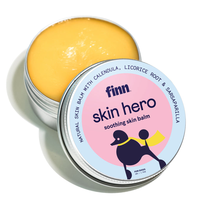 An open tin of Finn Skin Hero soothing skin balm for dogs, showing the yellow balm inside. The lid features a colorful label with a cartoon dog and text about natural ingredients like calendula and licorice root.