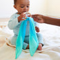 A baby in a light blue outfit eagerly reaches for the Silk and FSC-Certified Wood Baby Teether by Sarahs Silks as an adult offers it. The non-toxic teether, adorned with blue fabric, matches the cozy setting on a soft, light-colored blanket in the warm, brightly lit room.