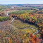 A winding river cuts through a forest of vibrant autumn foliage, with trees in shades of red, orange, yellow, and green, stretching toward distant hills and a lake under a partly cloudy sky.