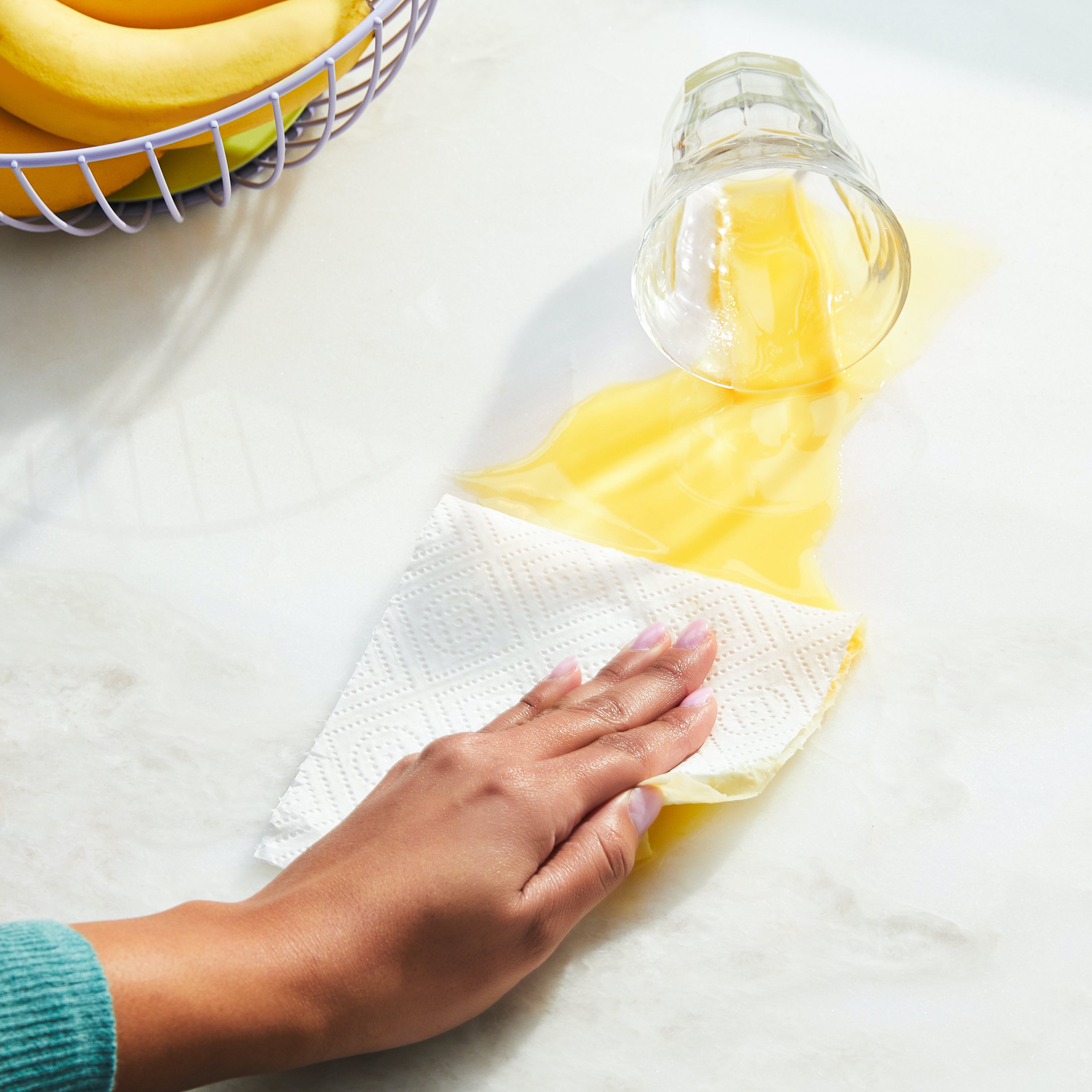 A hand uses Grove Co. Bamboo Paper Towels (140 sheets per roll) to clean up spilled orange juice on a white countertop, with a glass on its side and bananas in the background.