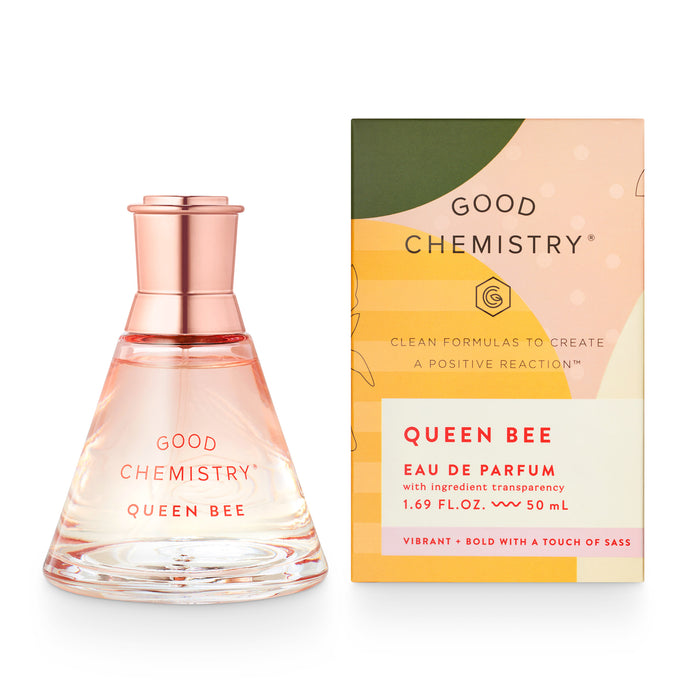 A bottle of Good Chemistrys Queen Bee perfume sits next to its box. The bottle is clear with a rose gold cap and pink text. The box is peach and green with product details and a bold, vibrant design.