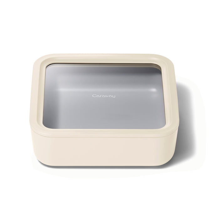 A square, cream-colored food storage container with rounded edges, a transparent lid, and the word Caraway printed in white on the gray interior bottom.