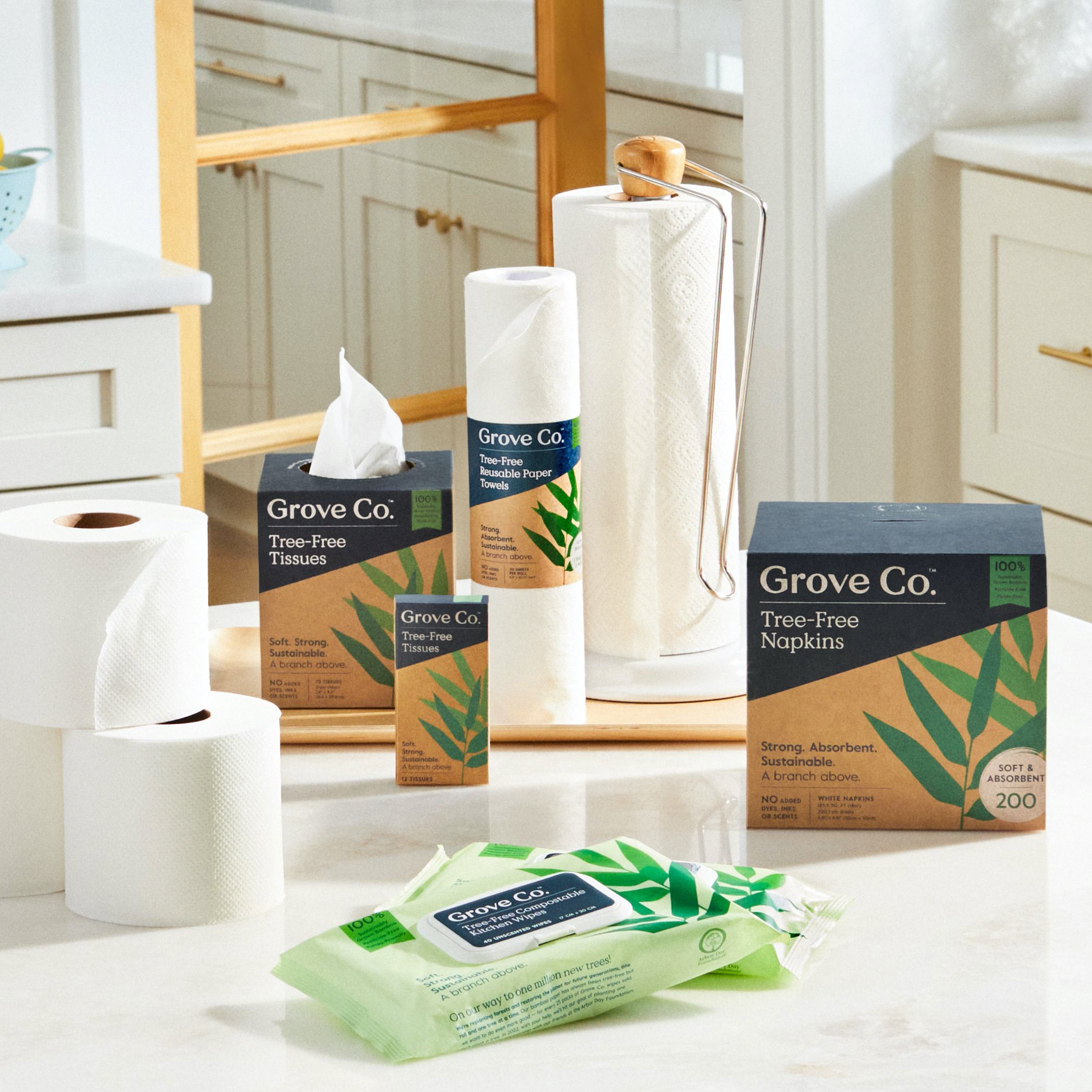 A kitchen countertop displays eco-friendly Grove Co. products, including tissues, tree-free toilet paper rolls, sustainable bamboo paper towels on a holder, napkins, and Tree-Free Compostable Kitchen Wipes made from 100% bamboo—all with green leaf designs.