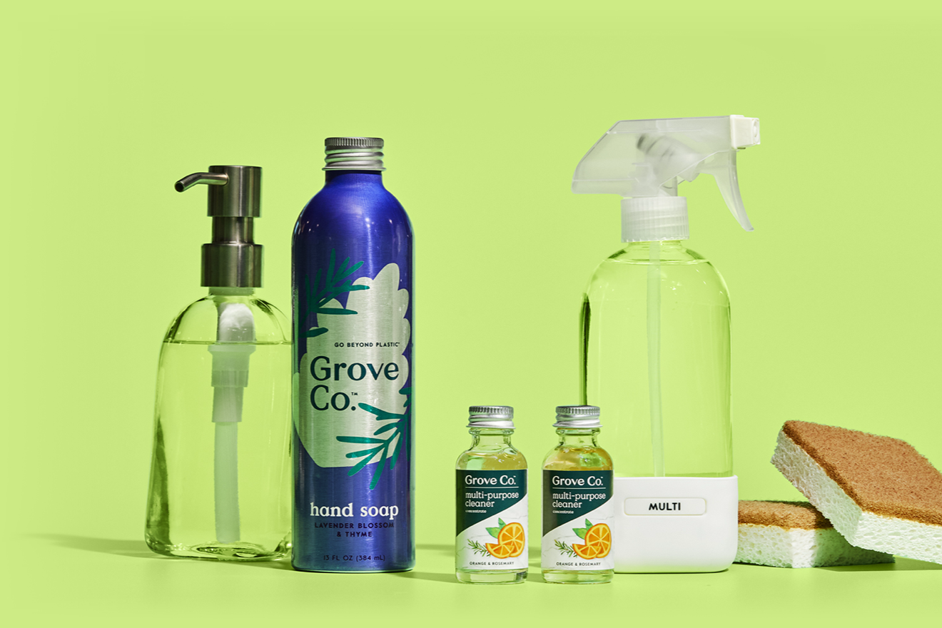 A set of eco-friendly cleaning supplies on a green background, including a glass soap dispenser, a Grove Co. hand soap bottle, two small bottles of cleaner, a spray bottle, and two scrub sponges.