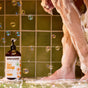 A person showers, covered in suds, with bubbles floating around. A bottle of Everyone 3-in-1 soap sits on green tiles near their feet against a tiled wall.