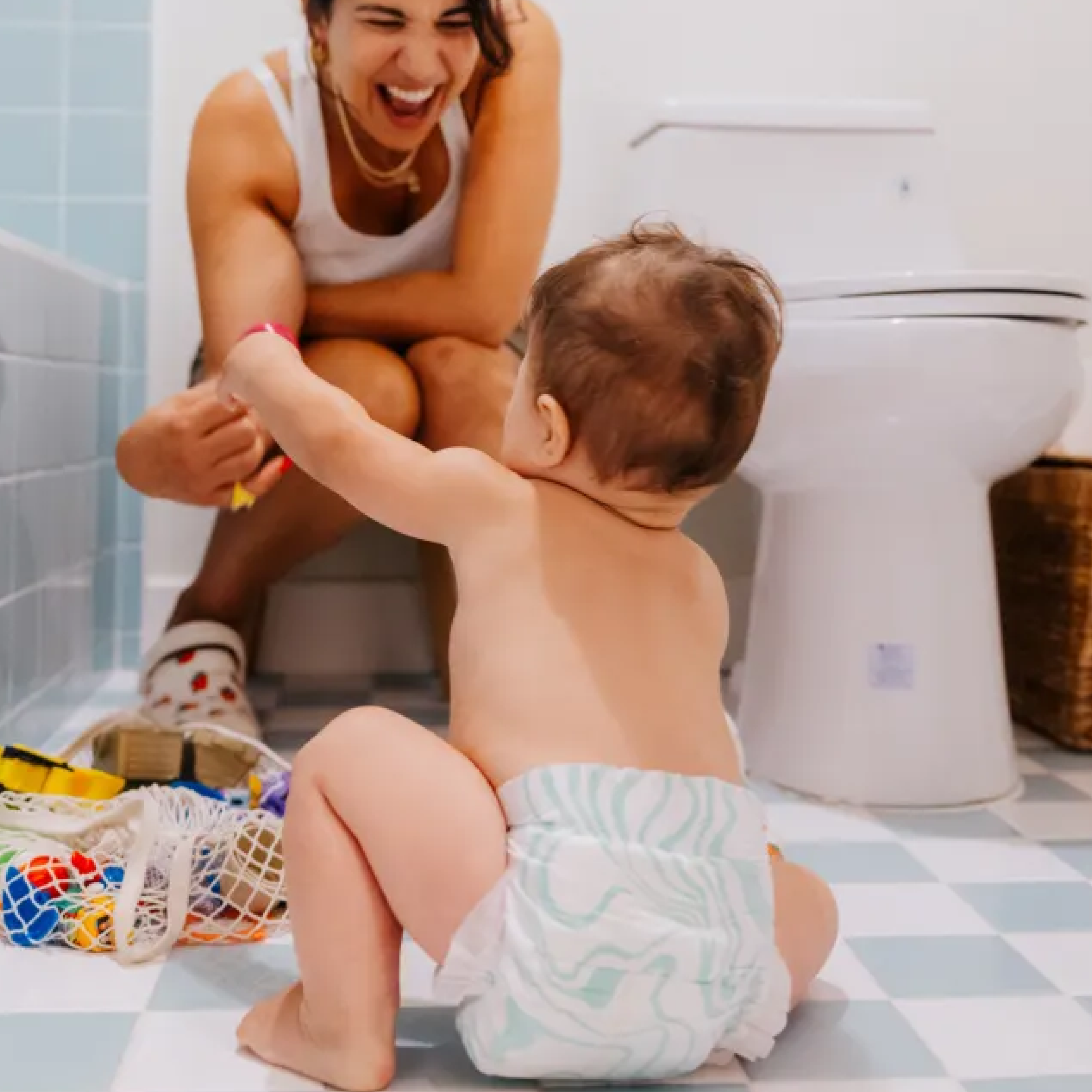 A smiling woman sits near a toilet, reaching out to a baby in a diaper who is squatting on a checkered bathroom floor, surrounded by colorful bath toys.