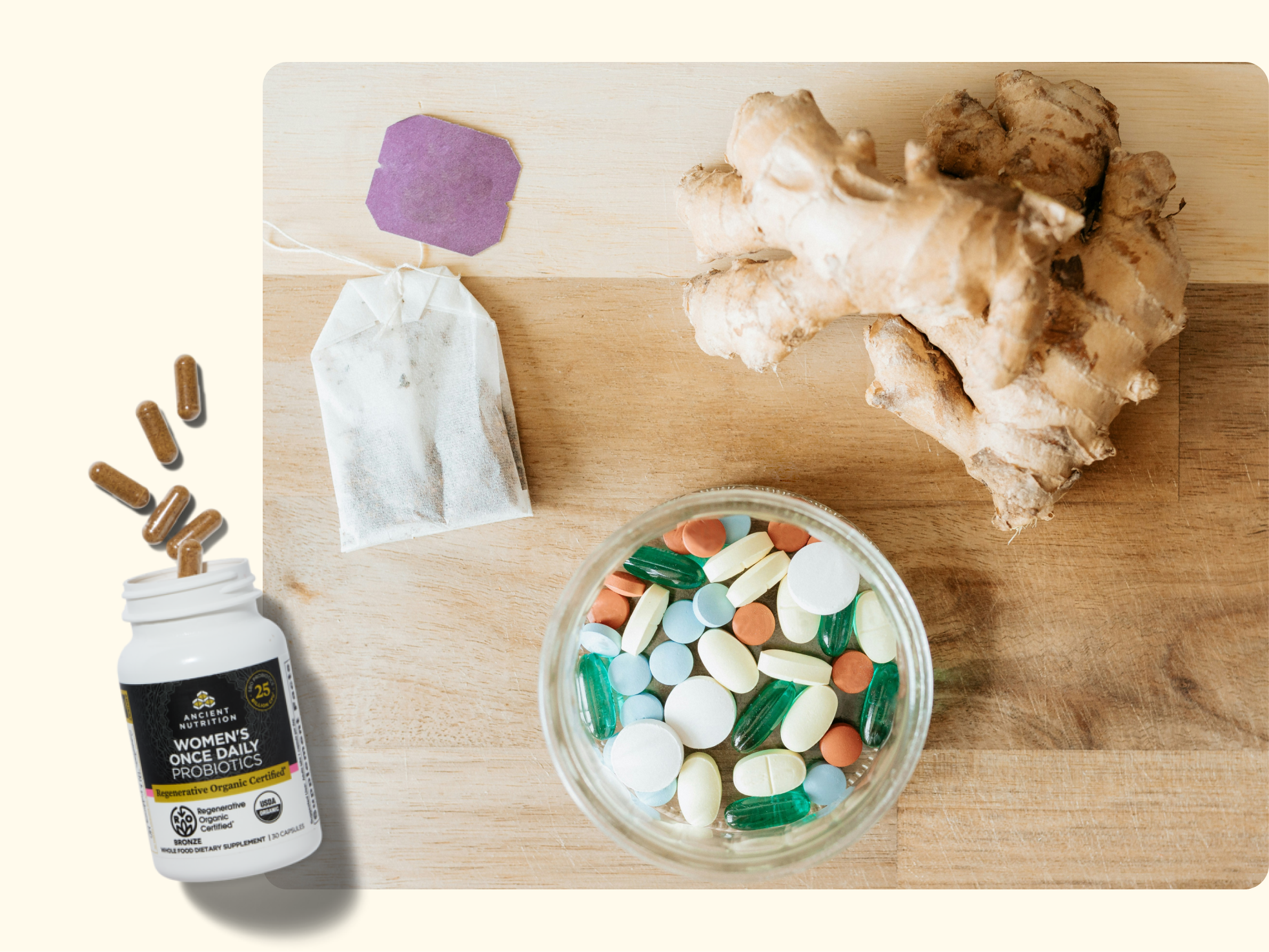 A wooden surface with ginger root, a jar filled with assorted pills and capsules, two tea bags (one purple, one white), and a bottle of women’s probiotics with capsules spilling out.