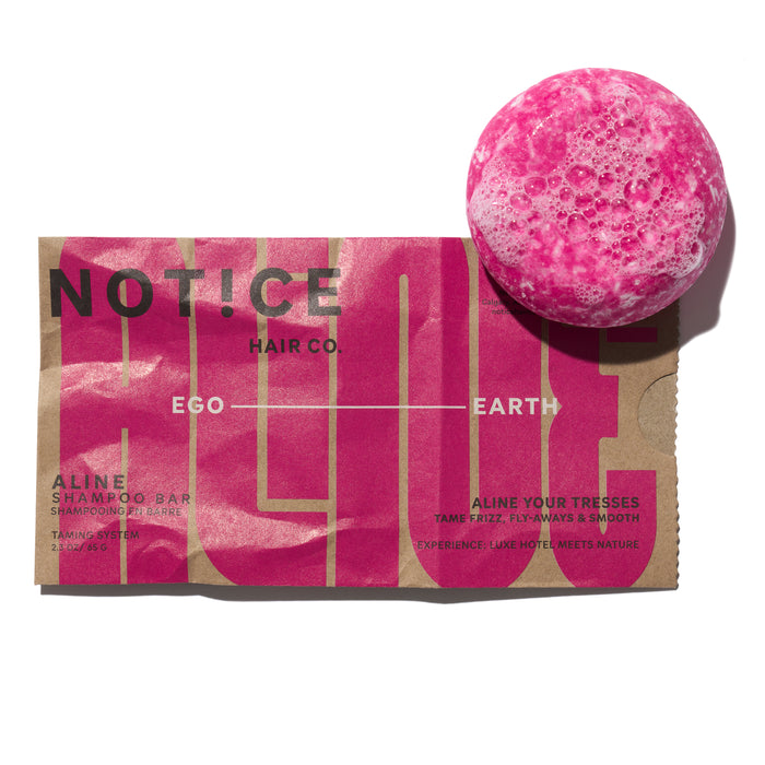 A bright pink, round shampoo bar sits above its brown and pink packaging labeled “NOT!CE HAIR CO.” The packaging features product details and bold text, partially covered by the shampoo bar.