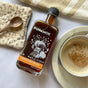 A bottle of Runamok Infused Maple Syrup - Pumpkin Spice sits beside a coffee cup, wooden spoon, patterned cloth, and striped napkin on a white surface.