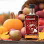 A bottle of Runamok Infused Maple Syrup - Pumpkin Spice sits among apples, pumpkins, and autumn leaves, with a tipped apple basket and a blurred fall landscape in the background.