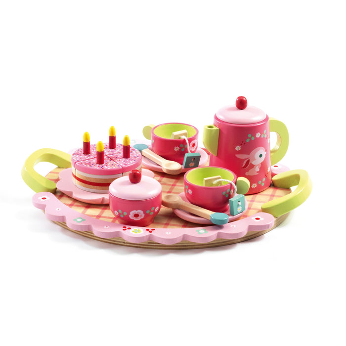 A colorful wooden tea set on a round tray with handles, featuring two cups, a teapot, a sugar bowl, spoons, a pink birthday cake with candles, and painted flower and bunny designs.