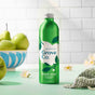A green metal bottle of Grove Co. apple & pear blossom hand soap sits on a light green surface, surrounded by fresh pears, white flowers, and a striped towel, with a white tiled wall in the background.