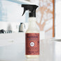 A bottle of Mrs. Meyers Multi-Surface Cleaner rests on a white kitchen counter, showcasing its cruelty-free formula suitable for any surface. In the background, a modern kitchen features a kettle and a blurred view through the window, creating an inviting atmosphere.