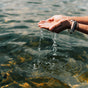 A person’s cupped hands lift water from a lake, with droplets falling back. The person wears several bracelets, and the sun reflects off the rippling surface of the water.