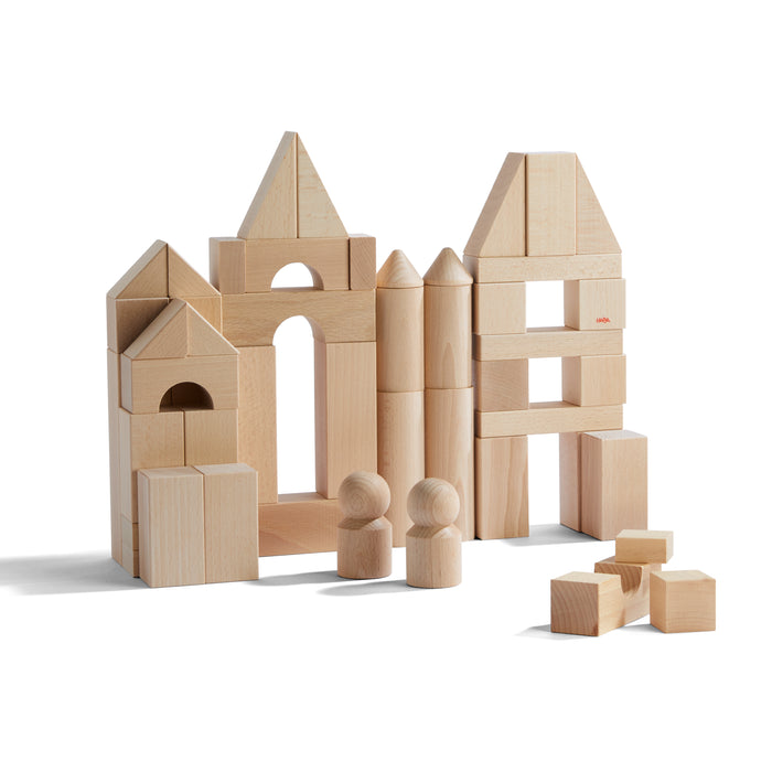 Wooden building blocks of various shapes are stacked to form structures resembling houses or castles. Two simple wooden human figures stand in front, with extra blocks scattered nearby. The background is plain white.