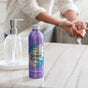 A purple aluminum bottle of Grove Co. lavender-scented hand soap stands on a marble counter next to a clear soap dispenser, while a person washes their hands at a sink in the background.