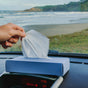 A hand pulls a cloth tissue from a blue LastTissue box on a car dashboard, with a scenic beach and ocean visible through the windshield in the background.