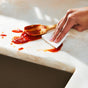 A hand wipes tomato sauce from a light-colored marble countertop with a white cloth. A wooden spoon covered in sauce rests on the countertop nearby.
