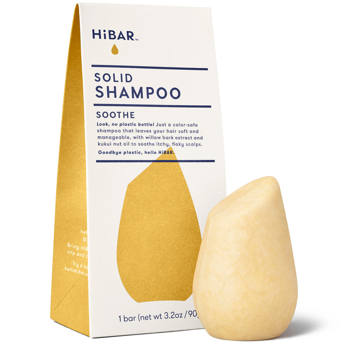 A bar of HiBAR solid shampoo stands in front of its triangular cardboard packaging. The packaging is cream and gold, displaying the product name and details. The shampoo bar is light yellow and teardrop-shaped.