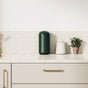 A clean, modern kitchen counter with a dark green canister, a white jar, and a small potted plant on a speckled countertop. Cream cabinets and gold hardware are visible below.