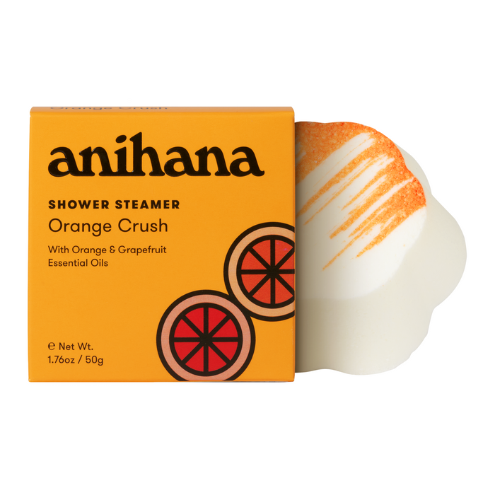A box labeled anihana Shower Steamer Orange Crush with orange and grapefruit graphics. Next to it is a white, round shower steamer with orange powder and drizzle on top.