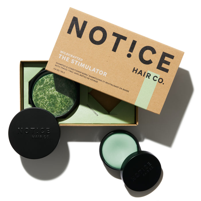 A box labeled NOTICE HAIR CO. THE STIMULATOR sits open, revealing a round green shampoo or conditioner bar. Two black-lidded containers, one open to show a light green product, are beside the box on a white background.
