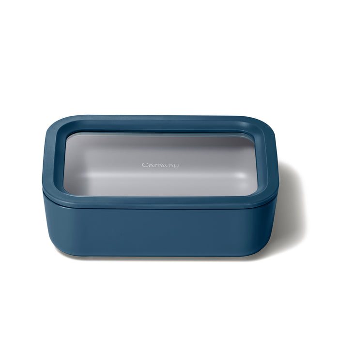A rectangular, dark blue food storage container with a transparent lid displaying the Caraway logo, pictured on a white background.