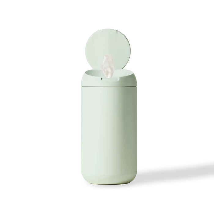 A light green cylindrical container with a hinged lid open, revealing a single white wipe being dispensed, set against a plain white background.