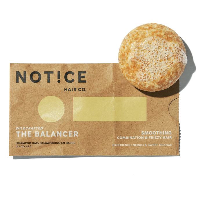 A round, foamy shampoo bar rests on brown packaging labeled NOTICE Hair Co. The Balancer, designed for smoothing combination and frizzy hair, with notes of neroli and sweet orange.